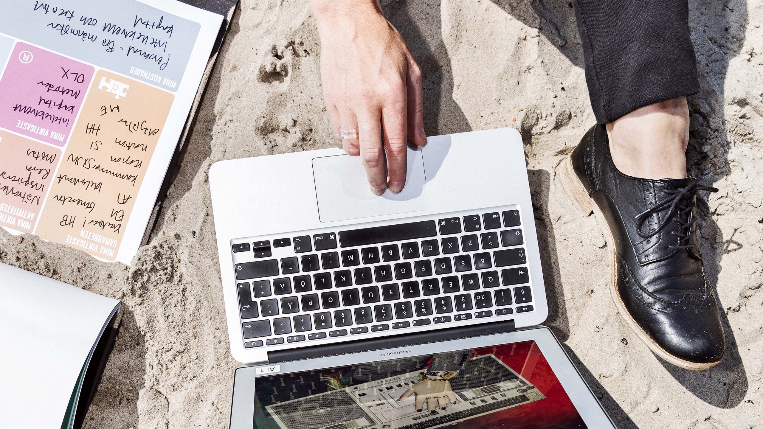 Person sitter med finkläder i sanden och trycker på tangentbordet till sin laptop.