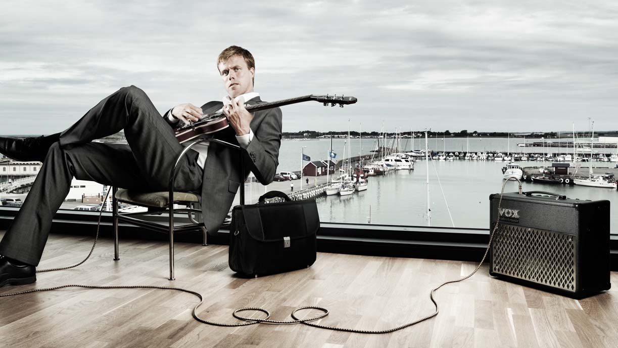 Person sitter med elgitarr vid ett panoramafönster över en hamn. Gitarren är kopplad till en högtalare.