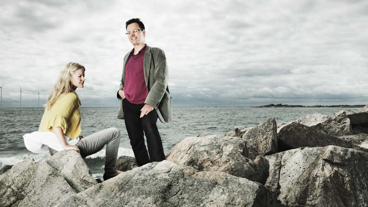 Två personer står vid större stenblock jämte ett hav. I bakgrunden syns en grå himmel.