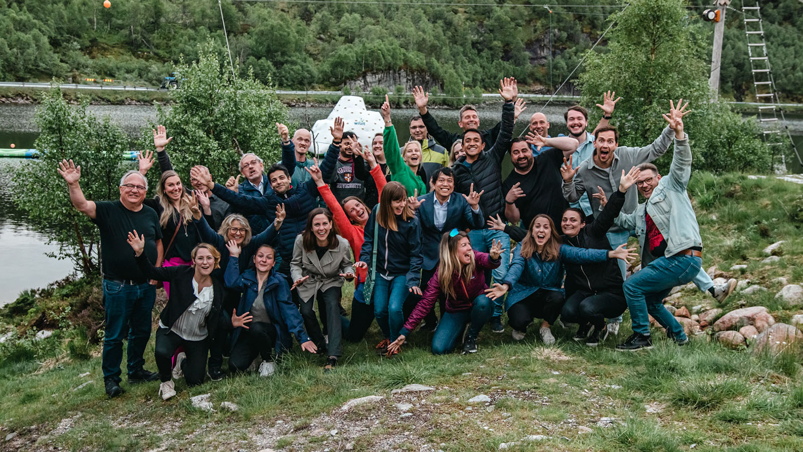 En grupp med människor står med armarna utsträckta och ler mot kameran, grönskande natur i bakgrunden.