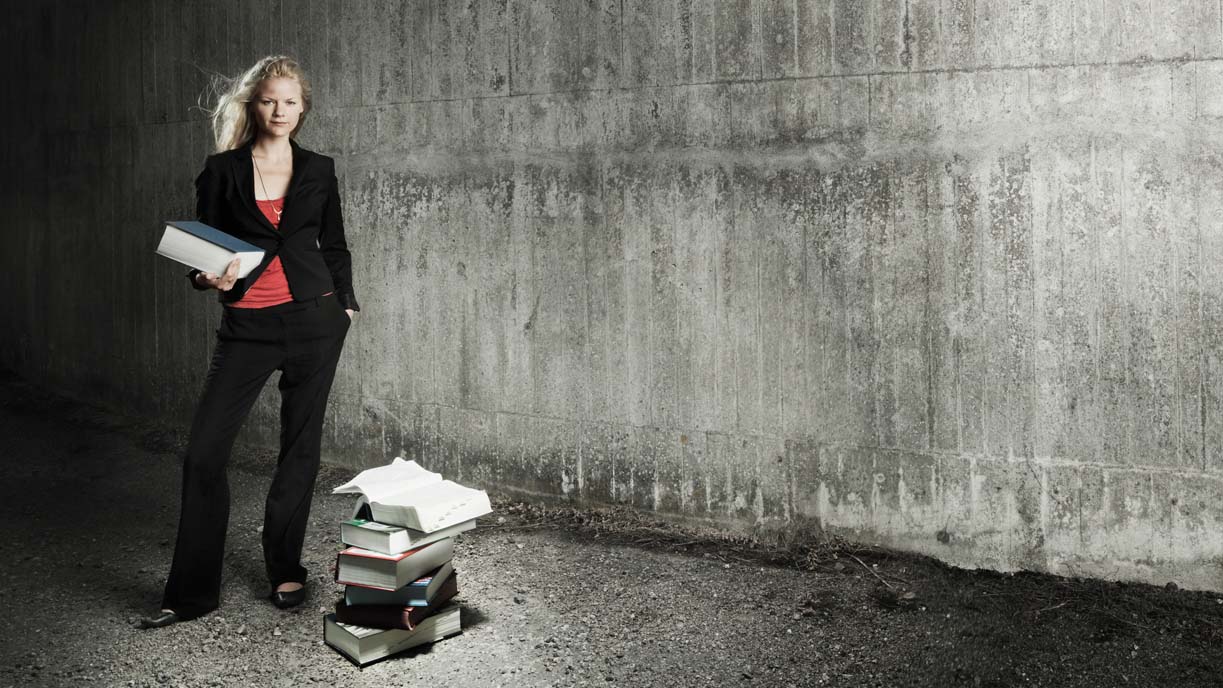 Person iklädd kavaj står vid en betongvägg med en lagbok i famnen. Fler stora, tunga böcker ligger på marken ijämte.