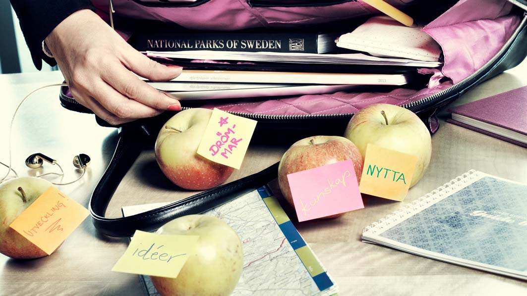 En väska innehållande böcker och anteckningar. Från väskan har äpplen och ytterligare anteckningar ramlat ner på ett bord.